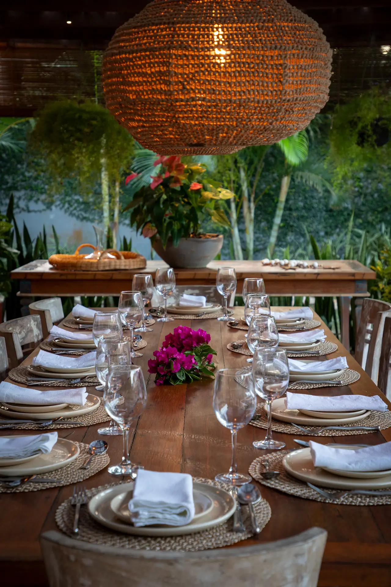 Mesa posta com pratos, taças e flores sob luminária de palha