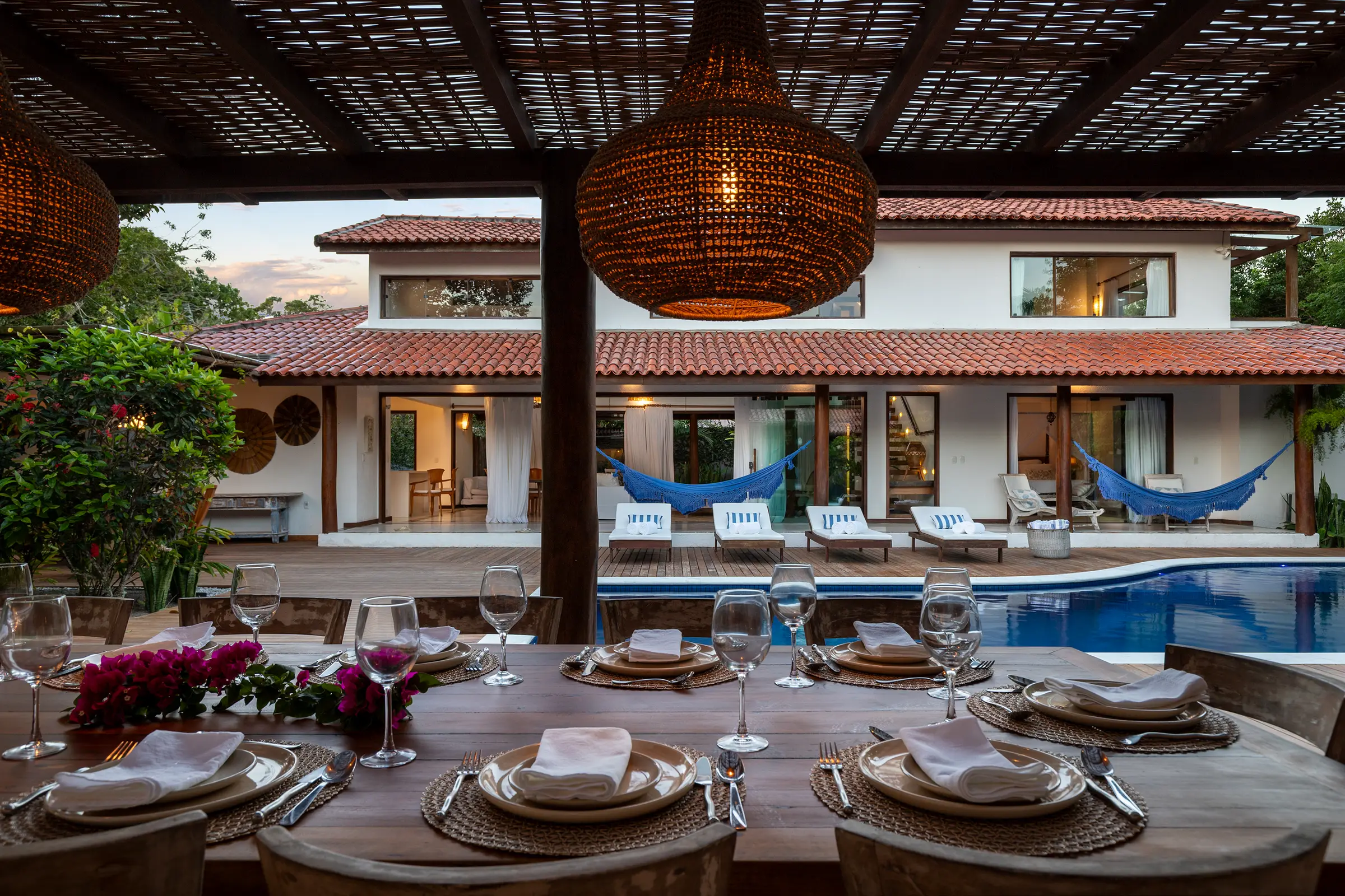 Mesa posta sob luminária de palha, com a casa e a piscina ao fundo