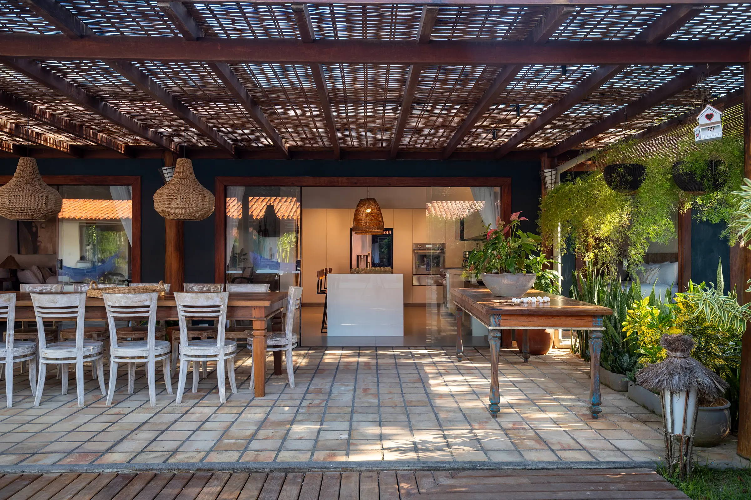 Terraço coberto com mesa comprida, cadeiras brancas e a cozinha ao fundo