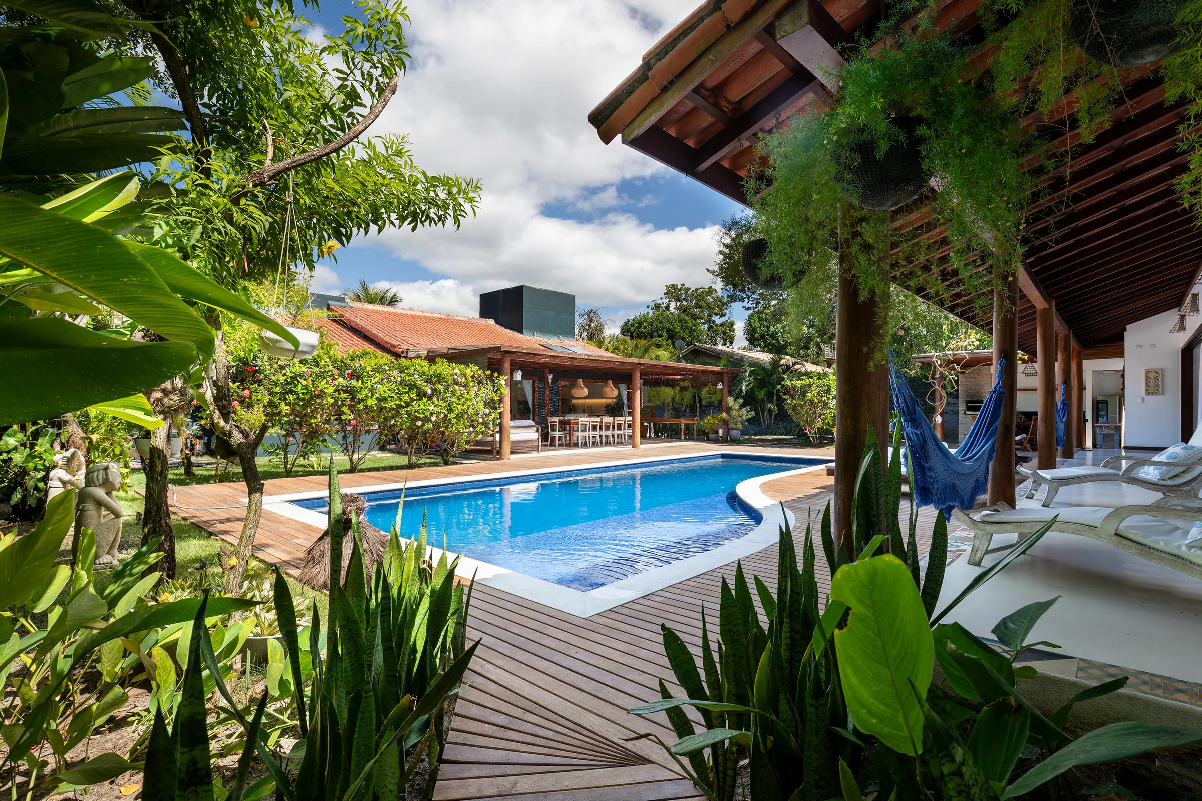 Piscina em curva entre jardim tropical e o beiral de madeira da casa, com céu azul