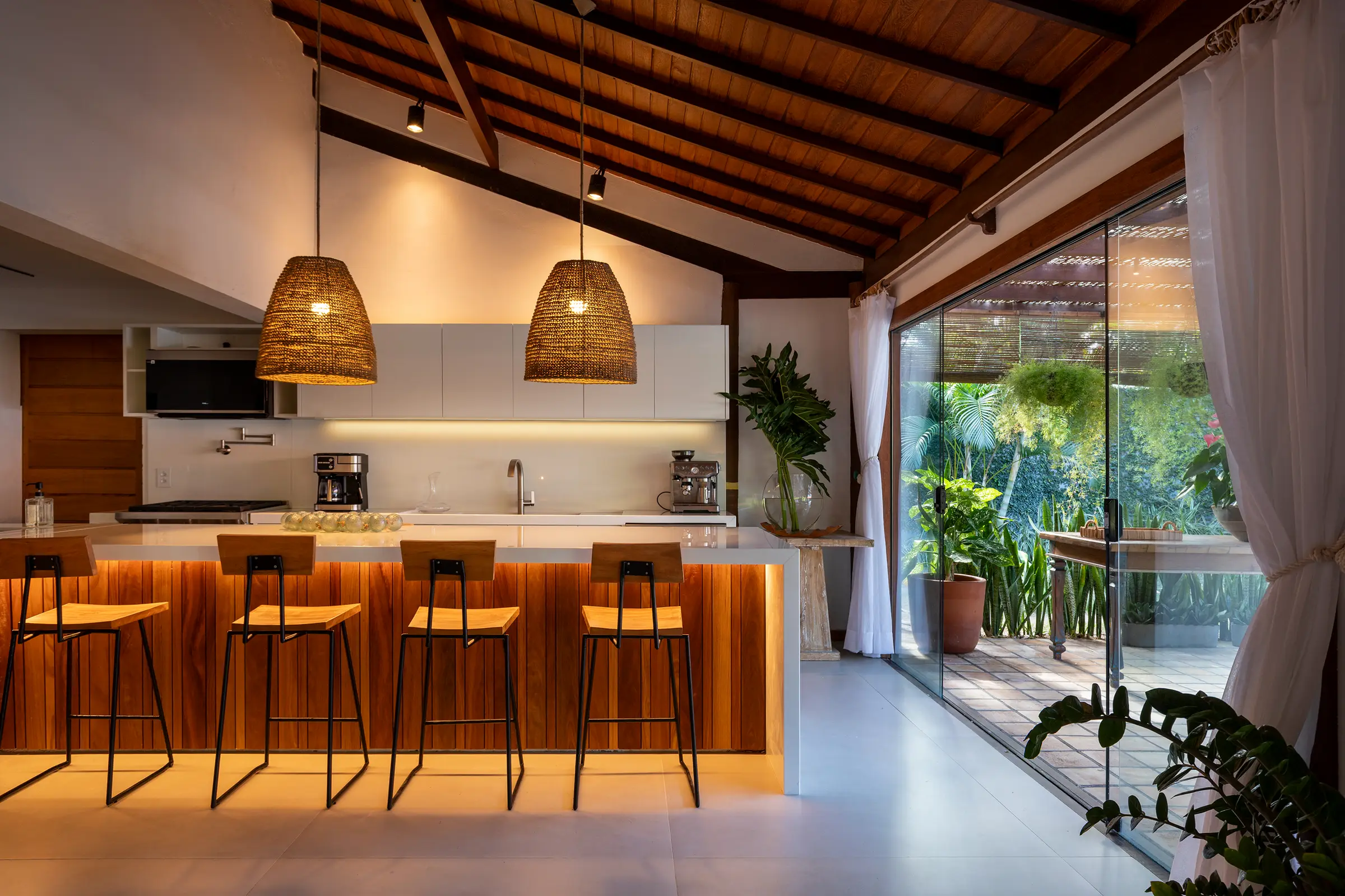 Cozinha com bancada de madeira iluminada, banquetas e janela para o jardim