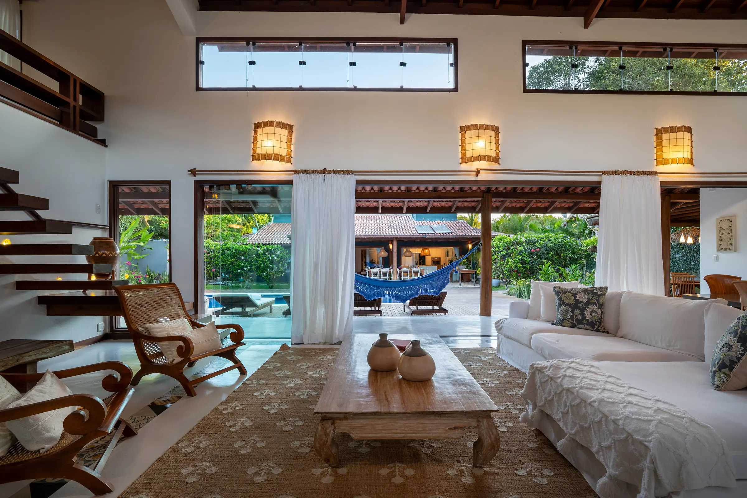 Sala com sofás brancos, escada de madeira e portas abertas para a piscina