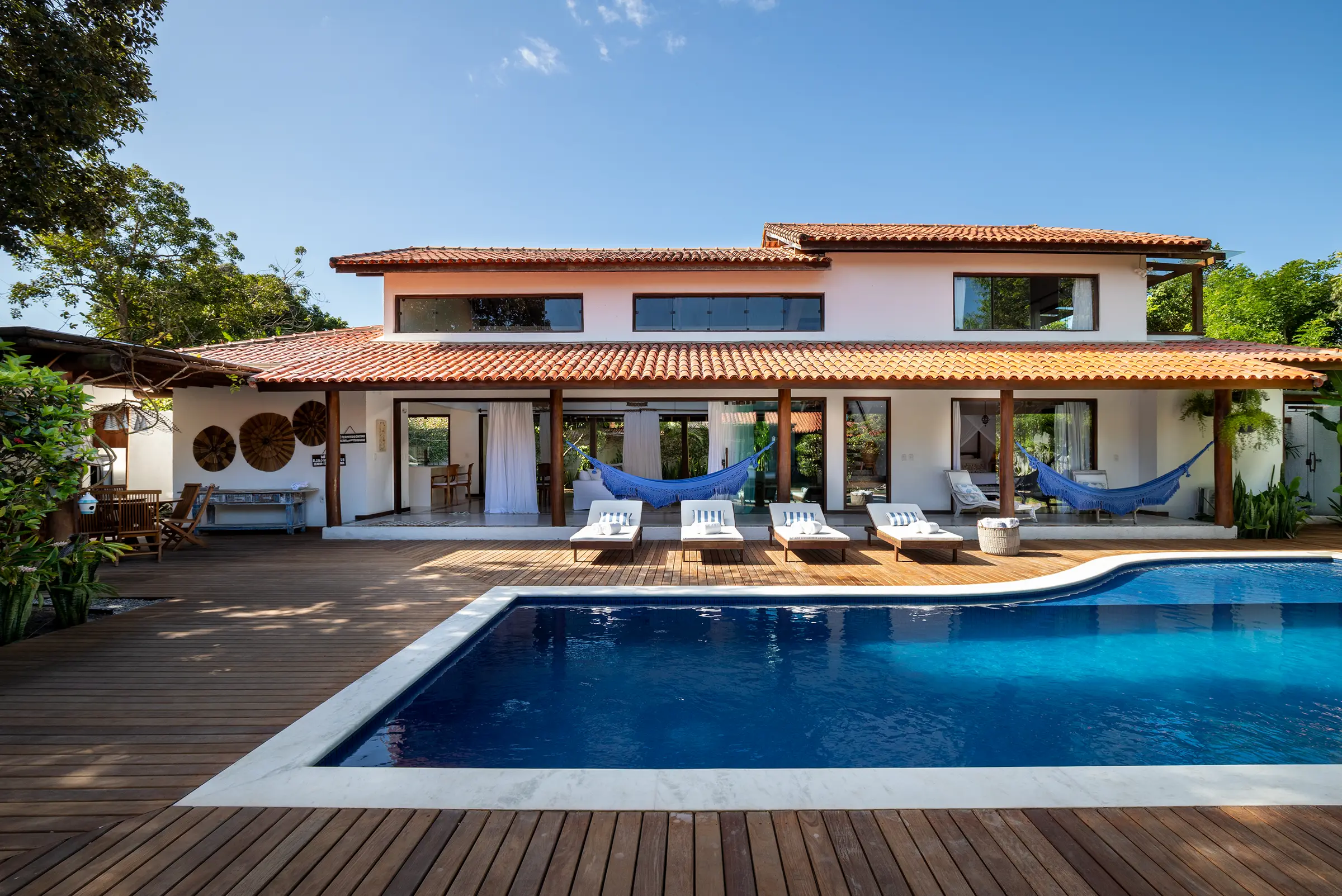 Fachada da casa com telhado de cerâmica, redes azuis e piscina em primeiro plano